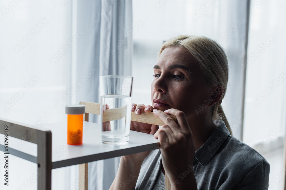 blonde woman with menopause looking at medication and glass of water on table. blonde woman with menopause looking at medication and glass of water on table.