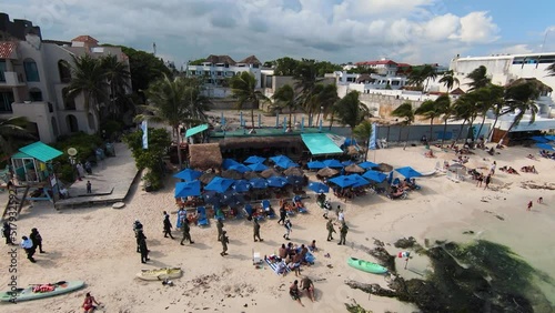 Drone clip above a busy Mexican beach resort with blue parasols and sun loungers in Playa del Carmen. Armed police officers patrol tourist area.
