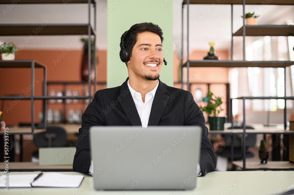 Smiling young hispanic salesman wearing headset is holding online call, male customer service representative sitting in front of a laptop and looking away
