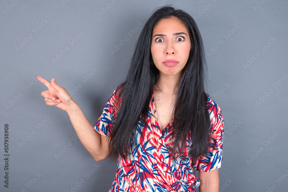 Serious Young latin woman wearing colorful dress over grey backgrogund smirks face points away on copy space shows something unpleasant. Look at this advertisement. Big price concept Serious Young latin woman wearing colorful dress over grey backgrogund smirks face points away on copy space shows something unpleasant. Look at this advertisement. Big price concept