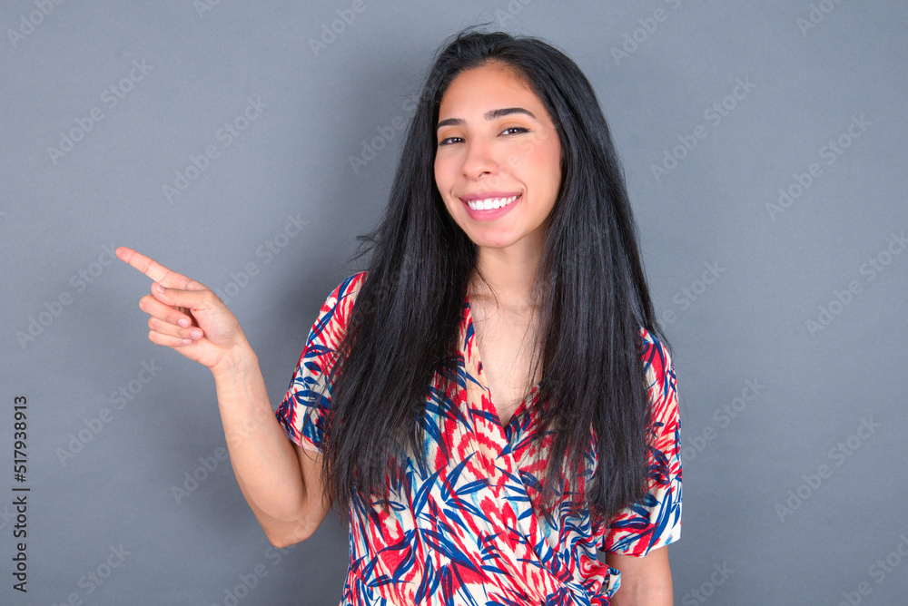 Young latin woman wearing colorful dress over grey backgrogund points to side on blank space demonstrates advertisement. People and promotion concept Young latin woman wearing colorful dress over grey backgrogund points to side on blank space demonstrates advertisement. People and promotion concept
