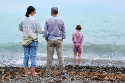 Three people are standing on the seashore. One man and two women are resting at the water's edge, watching the waves crash on the shore. A woman in shorts wets her feet in the sea. The couple is talki photo