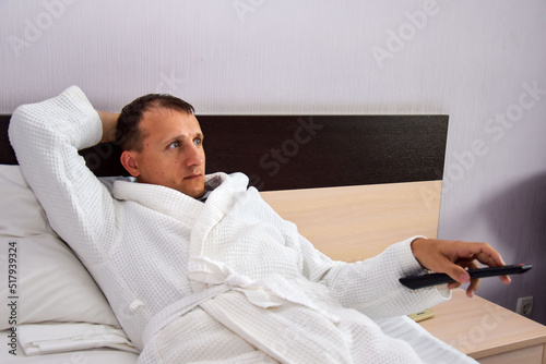 A man lies on a bed in a bathrobe, holds a remote control in his hand and switches the channel. Lonely man on the bed watching TV.