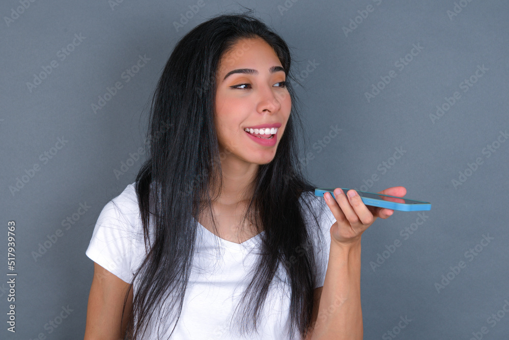Smiling young beautiful brunette woman wearing white t-shirt over grey background sending voice message on her smart phone. Communication and new technologies concept. Smiling young beautiful brunette woman wearing white t-shirt over grey background sending voice message on her smart phone. Communication and new technologies concept.