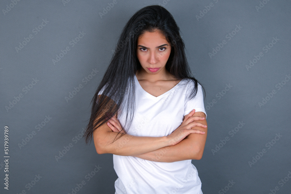 Picture of angry young beautiful brunette woman wearing white t-shirt over grey background crossing arms. Looking at camera with disappointed expression. Picture of angry young beautiful brunette woman wearing white t-shirt over grey background crossing arms. Looking at camera with disappointed expression.