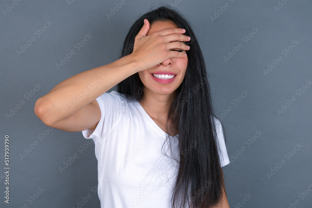 Happy young beautiful brunette woman wearing white t-shirt over grey wall closing eyes with hand going to see surprise prepared by friend standing and smiling in anticipation for something wonderful. Happy young beautiful brunette woman wearing white t-shirt over grey wall closing eyes with hand going to see surprise prepared by friend standing and smiling in anticipation for something wonderful.