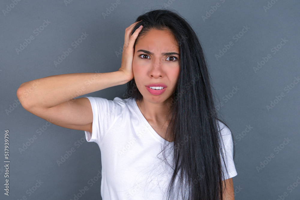 Obraz premium Portrait of confused young beautiful brunette woman wearing white t-shirt over grey wall holding hand on hair and frowning, panicking, losing memory. Worried and anxious can not remember anything.