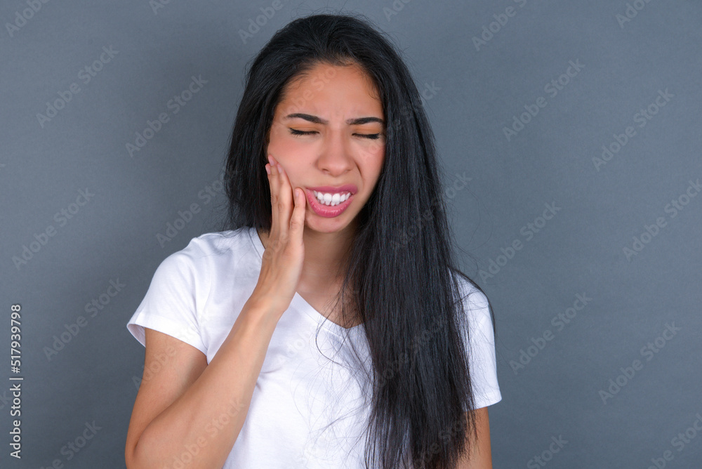 Tooth ache concept. young beautiful brunette woman wearing white t-shirt over grey feeling pain, holding his cheek with hand, suffering from bad toothache, looking at camera with painful expression Tooth ache concept. young beautiful brunette woman wearing white t-shirt over grey feeling pain, holding his cheek with hand, suffering from bad toothache, looking at camera with painful expression
