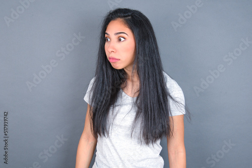 young beautiful brunette woman wearing white t-shirt over grey background stares aside with wondered expression has speechless expression. Embarrassed model looks in surprise