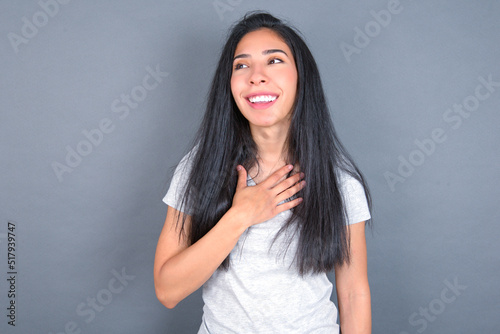 Joyful young beautiful brunette woman wearing white t-shirt over grey background expresses positive emotions recalls something funny keeps hand on chest and giggles happily.