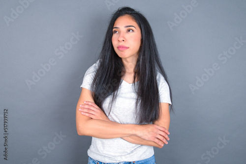 Charming thoughtful young beautiful brunette woman wearing white t-shirt over grey background stands with arms folded concentrated somewhere with pensive expression thinks what to do