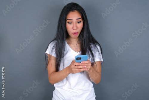 young beautiful brunette woman wearing white t-shirt over grey background looks with bugged eyes, holds modern smart phone, receives unexpected message from friend, reads reminder.