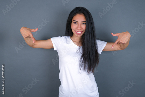 Pick me! Confident, self-assured and charismatic young beautiful brunette woman wearing white t-shirt over grey background promoting oneself as wanting role smiling broadly and pointing at body.