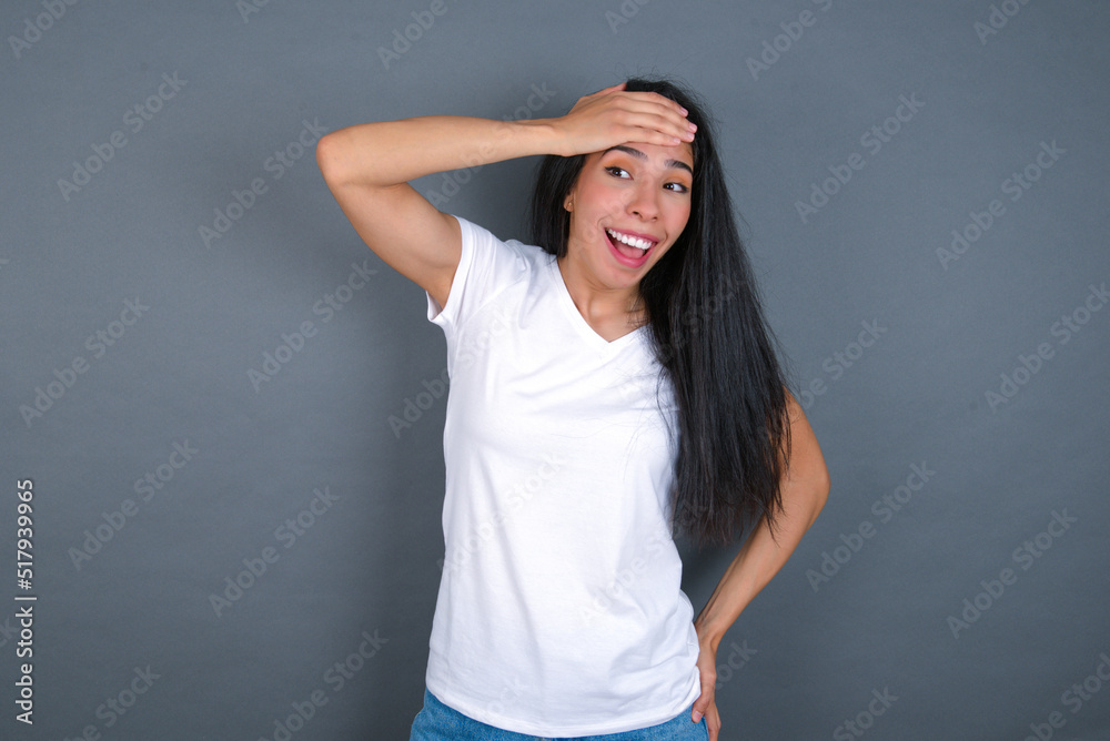 young beautiful brunette woman wearing white t-shirt over grey background touching forehead, hears something surprising, glad receive good news, feels relieved. Almost got in trouble. young beautiful brunette woman wearing white t-shirt over grey background touching forehead, hears something surprising, glad receive good news, feels relieved. Almost got in trouble.