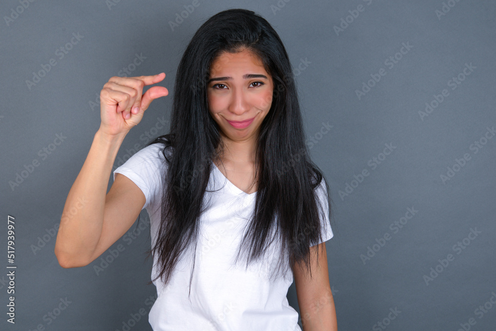 Upset young beautiful brunette woman wearing white t-shirt over grey background shapes little gesture with hand demonstrates something very tiny small size. Not very much Upset young beautiful brunette woman wearing white t-shirt over grey background shapes little gesture with hand demonstrates something very tiny small size. Not very much