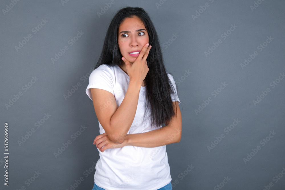 young beautiful brunette woman wearing white t-shirt over grey background covering mouth with hands scared from something or someone bitting nails young beautiful brunette woman wearing white t-shirt over grey background covering mouth with hands scared from something or someone bitting nails