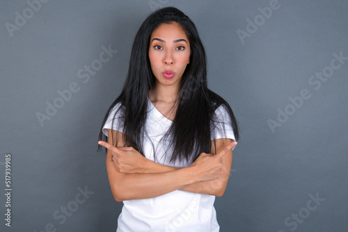 Confused young beautiful brunette woman wearing white t-shirt over grey background chooses between two ways, points at both sides with crossed hands, feels doubt. Need your advice.