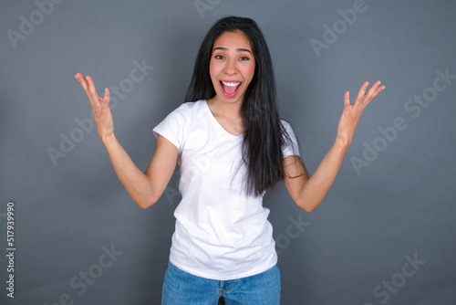 Joyful excited lucky young beautiful brunette woman wearing white t-shirt over grey background cheering, celebrating success, screaming yes with clenched fists