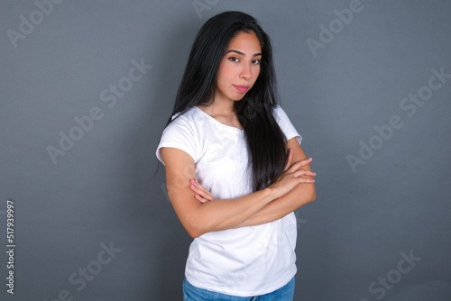 Waist up shot of self confident young beautiful brunette woman wearing white t-shirt over grey background has broad smile, crosses arms, happy to meet with colleagues.