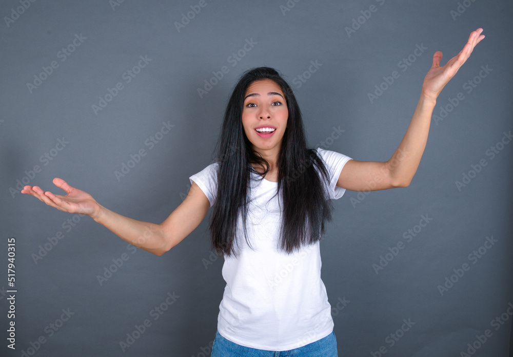 Cheerful young beautiful brunette woman wearing white t-shirt over grey background making a welcome gesture raising arms over head. Cheerful young beautiful brunette woman wearing white t-shirt over grey background making a welcome gesture raising arms over head.