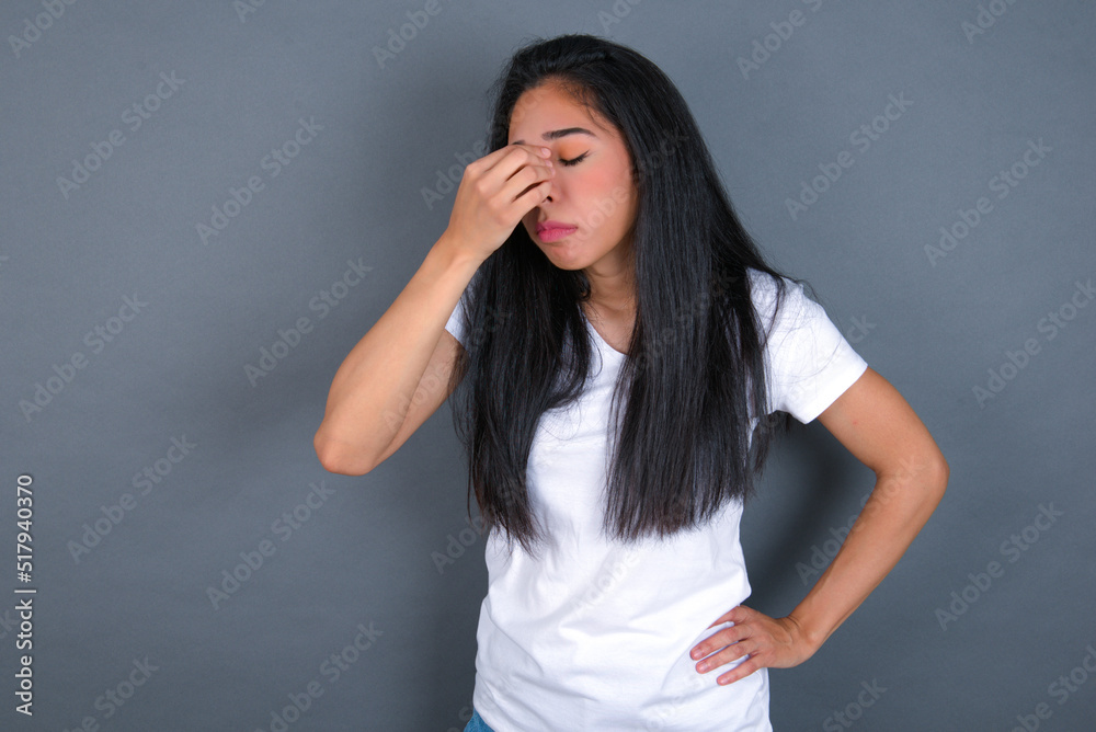 Very upset, young beautiful brunette woman wearing white t-shirt over grey background touching nose between closed eyes, wants to cry, having stressful relationship or having troubles with work Very upset, young beautiful brunette woman wearing white t-shirt over grey background touching nose between closed eyes, wants to cry, having stressful relationship or having troubles with work