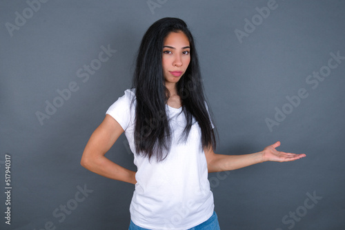 Portrait of young beautiful brunette woman wearing white t-shirt over grey background with arm out in a welcoming gesture.