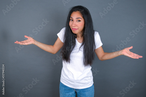 Papier peint Puzzled and clueless young beautiful brunette woman wearing white t-shirt over grey background with arms out, shrugging shoulders, saying: who cares, so what, I don't know