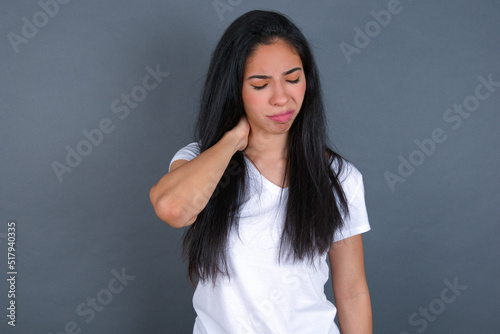 young beautiful brunette woman wearing white t-shirt over grey background suffering from back and neck ache injury, touching neck with hand, muscular pain.