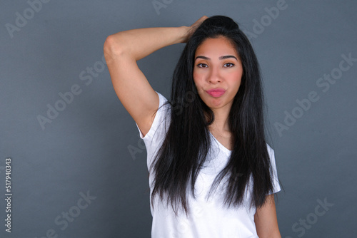 young beautiful brunette woman wearing white t-shirt over grey background saying: Oops, what did I do? Holding hand on head with frightened and regret expression.