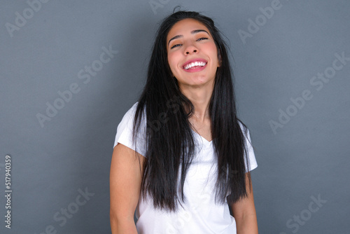 young beautiful brunette woman wearing white t-shirt over grey background with broad smile, shows white teeth, feeling confident rejoices having day off.