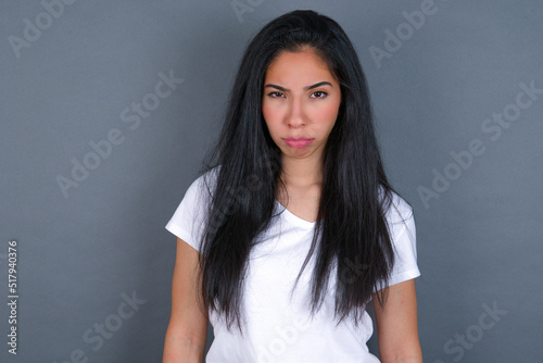 young beautiful brunette woman wearing white t-shirt over grey background frowning his eyebrows being displeased with something.