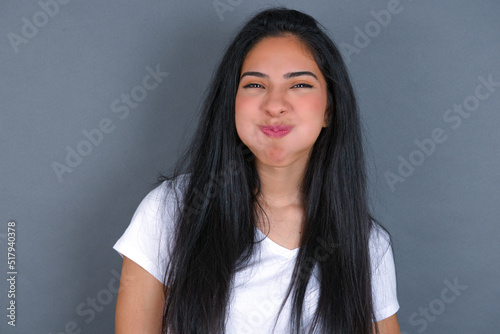 young beautiful brunette woman wearing white t-shirt over grey background puffing cheeks with funny face. Mouth inflated with air, crazy expression.