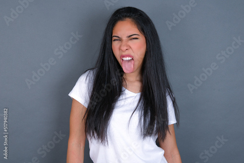 young beautiful brunette woman wearing white t-shirt over grey background with happy and funny face smiling and showing tongue.