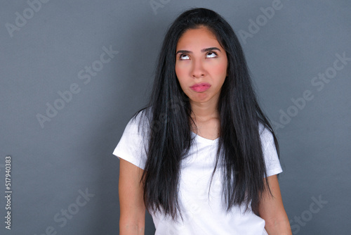 young beautiful brunette woman wearing white t-shirt over grey background has worried face looking up lips together, being upset thinking about something important, keeps hands down.