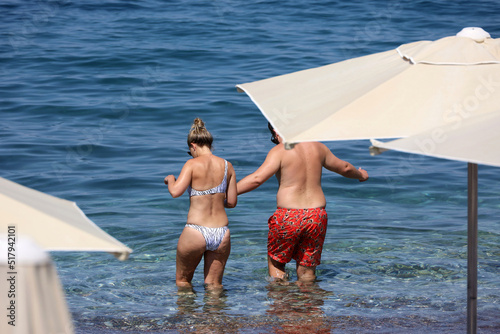 Fototapeta Naklejka Na Ścianę i Meble -  Couple going to swim in blue transparent water, view through sun umbrellas. Man and woman in swimsuit holding hands, vacation on summer beach
