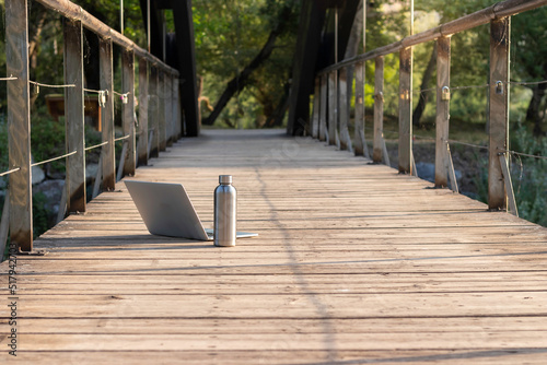 Fototapeta Naklejka Na Ścianę i Meble -  Isolated front view of laptop and metl bottle of water on a wooden flooring with nature in the background and copy space