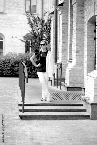 Black and white photo elegant business woman in sunglasses in a stylish black blouse and white trousers on a city street. Architecture and cityscape background. Sunny day. Tourism and travel concept