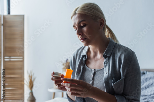 frustrated blonde woman with menopause holding bottle with pills and sitting in bedroom.