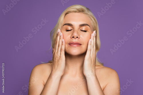 blonde woman with bare shoulders and closed eyes applying cream isolated on purple.