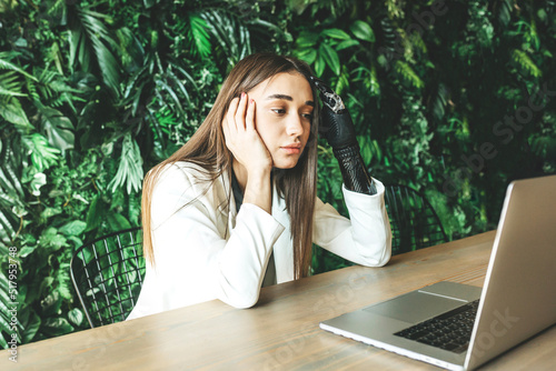 A tired young girl with a prosthetic bionic arm is studying or working on a laptop against a wall of plants. Online work, distance learning