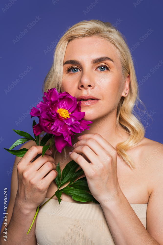 blonde woman with blue eyes holding blooming flower and looking at camera isolated on purple.
