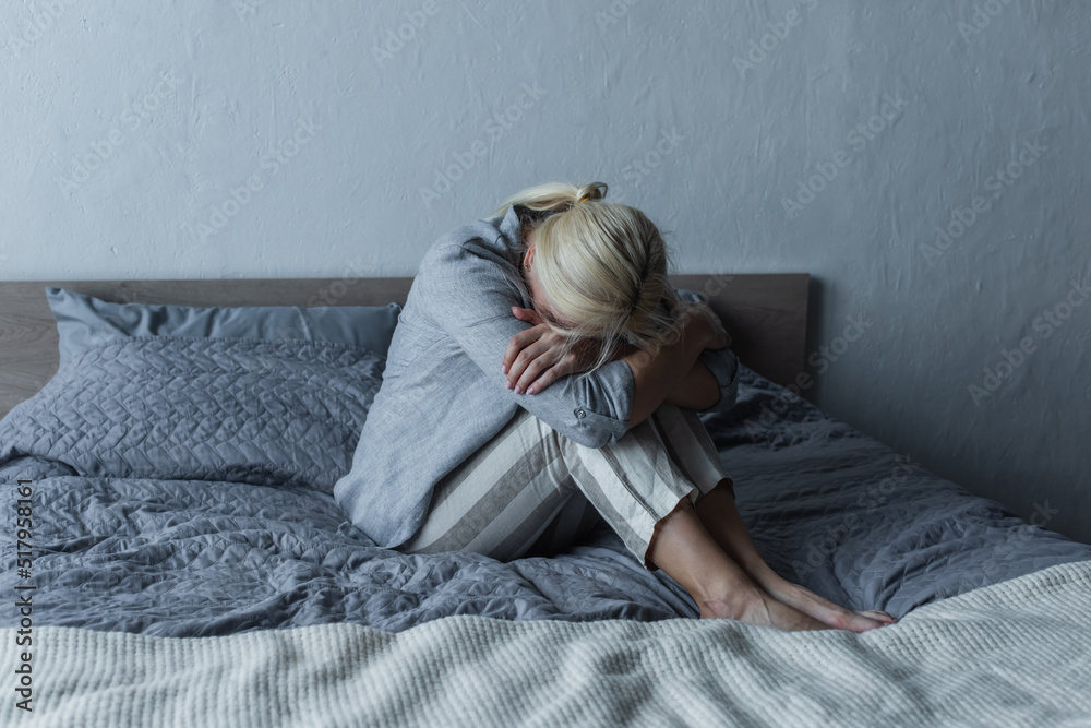 blonde woman covering face and hugging knees on bed while feeling pain in stomach during menopause. blonde woman covering face and hugging knees on bed while feeling pain in stomach during menopause.