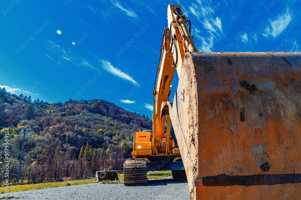 Close-up of a large bucket of a yellow excavator on a rubble square. Background of mountains, forest, blue sky. Low angle photo.