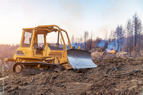 View of a bulldozer working in a forest on a slope. The bulldozer clears the slope. A fire in the forest. Smoke.
