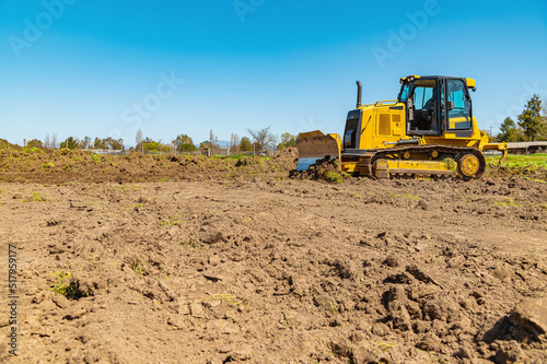 Sidet view of a yellow bulldozer on a plot of land. A bulldozer clears the area. Blue sky background.
