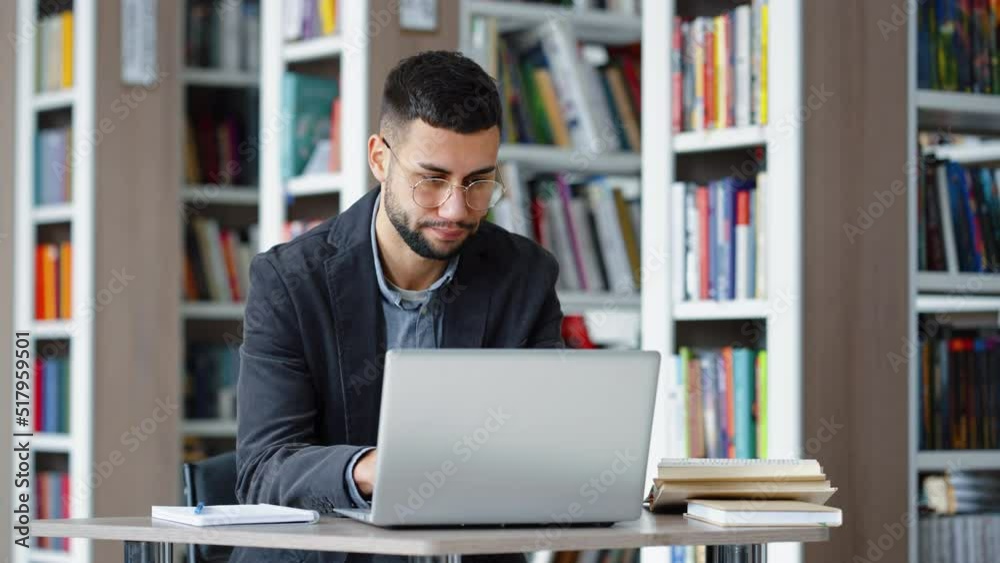 Handsome young man with eyeglasses sitting in library and browsing internet on laptop, looking stressed and unhappy. Distance learning. Remote work. Concept of technology Handsome young man with eyeglasses sitting in library and browsing internet on laptop, looking stressed and unhappy. Distance learning. Remote work. Concept of technology