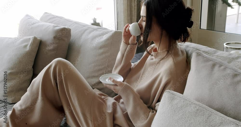 A beautiful Young woman is drinking coffee and tea in the living room at home, healthy calm lady relaxing on comfortable sofa feel stress free at home lounge alone. Woman rests at home on luxury sofa.