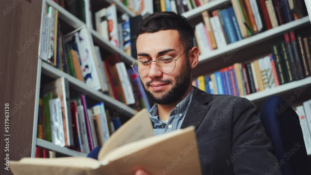 Cheerful young man with eyeglasses sitting by bookshelves and reading book, flipping pages. Low angle portrait of reader enjoying literature in book shop or library. Concept of education Cheerful young man with eyeglasses sitting by bookshelves and reading book, flipping pages. Low angle portrait of reader enjoying literature in book shop or library. Concept of education
