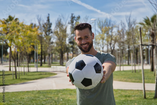 Young and handsome man, with a beard and green shirt, with blue eyes, perfect smile, offering a soccer ball, smiling. Concept sport, ball, football, world, competition. © Manuel
