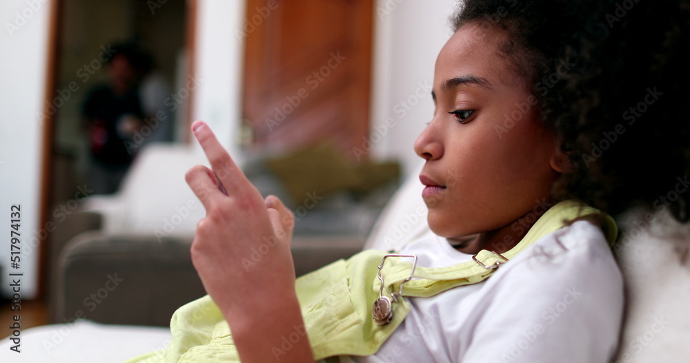 Obraz premium Little black girl using smartphone device at home sofa. African ethnicity